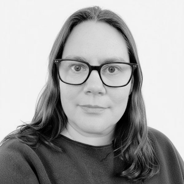 A black and white photograph of a white woman looking directly at the camera. She wears glasses and has long dark hair.
